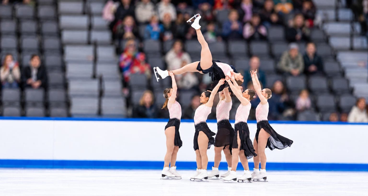 Teammates lifting a skater high above the ice in a group lift, her skate pointing to the ceiling
