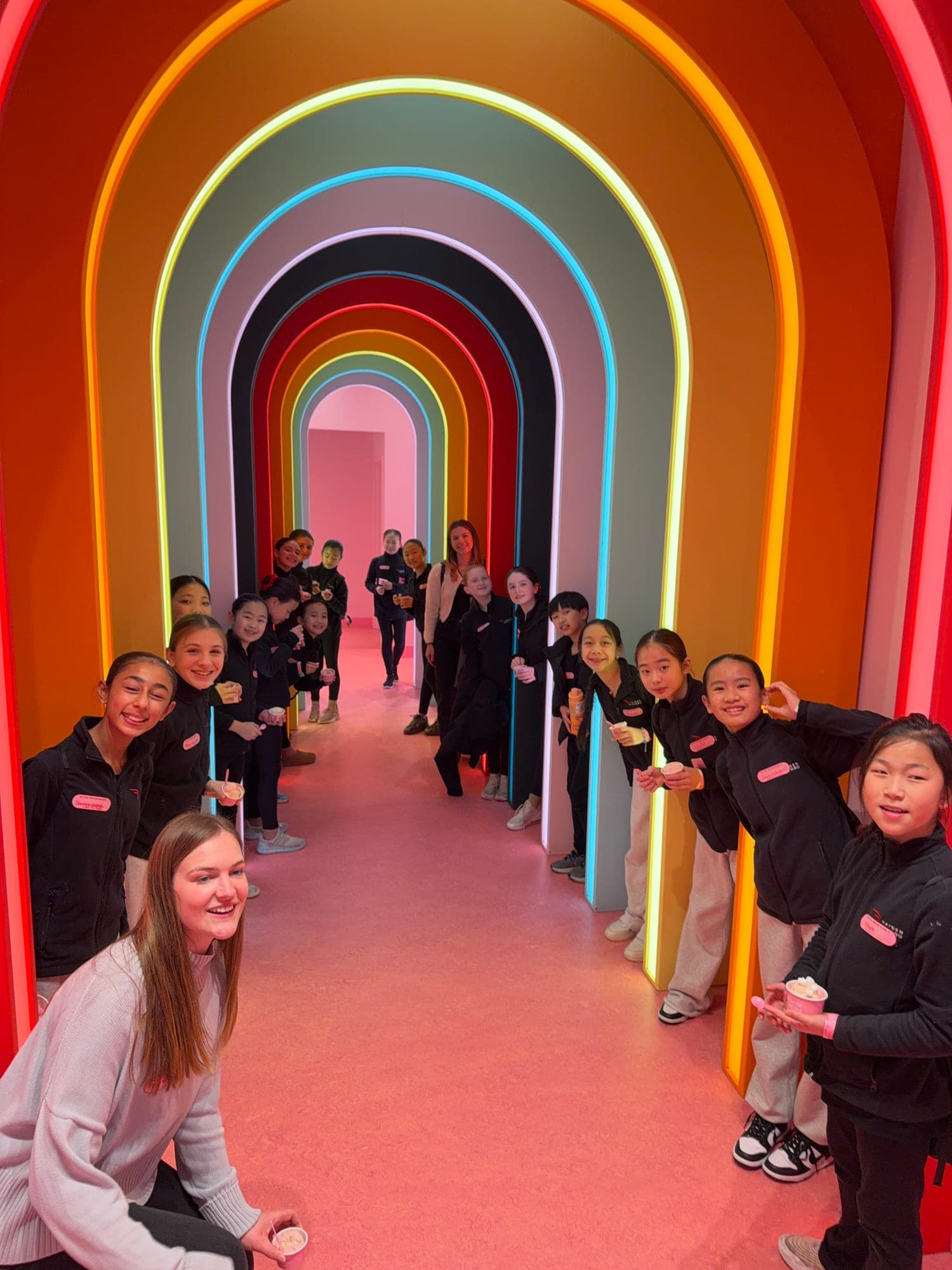 Young Hayden Synchro skaters and their coach smiling on a team outing, lined up along a rainbow tunnel